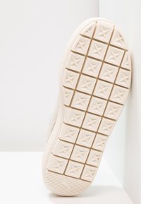 Semelle d'une basket beige avec des motifs carrés en relief pour l'adhérence et un logo Puma près du talon, positionnée contre un mur blanc en angle.
