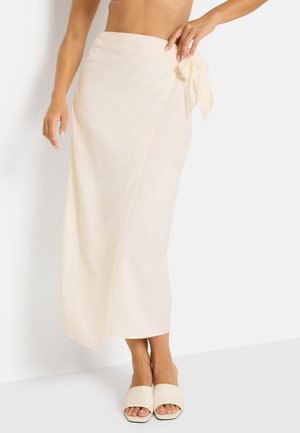 Woman wearing a cream wrap skirt tied at the waist and matching cream open-toe slide sandals against a white background.