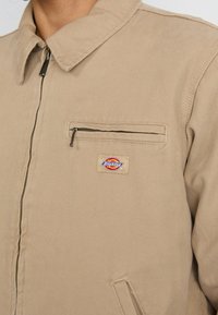 Beige work jacket in textured cotton fabric, featuring a collar, front zipper, and a small logo patch on the chest pocket.