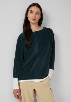 Haut en velours vert foncé avec un col rond, des manches trois-quarts et une coupe décontractée, superposé sur une chemise à manches longues blanche.