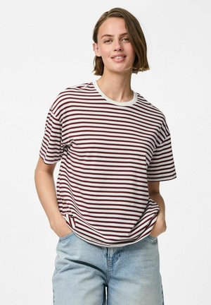 Ung kvinne med kort hår iført en rødbrun og hvit stripete t-skjorte og lyseblå jeans, stående med hendene i lommene og smilende.