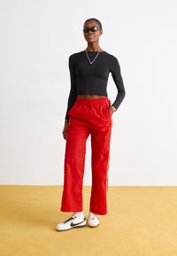 Črn dolg rokavni crop top v kombinaciji s sproščenimi rdečimi trenirkami z belimi stranskimi črtami. Obut z belimi in črnimi čevlji ter sončnimi očali.