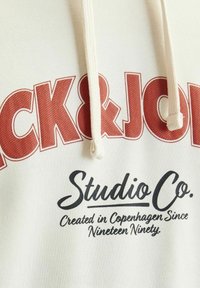 Sudadera de un beige claro con texto en negrita rojo y negro: "JACK & JONES" y "Studio Co. Creado en Copenhague desde mil novecientos noventa."