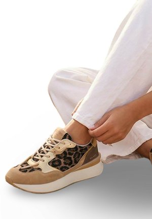 Personne portant un pantalon blanc qui attache des baskets beige imprimé léopard en étant assise devant un fond blanc.