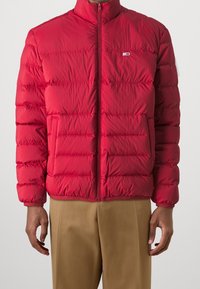 Homme portant une veste matelassée rouge vif avec fermeture éclair et un pantalon beige, se tenant avec les bras détendus sur les côtés contre un fond blanc.