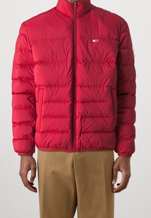 Mann in einer leuchtend roten Steppjacke mit Reißverschluss und beigen Hosen, stehend mit entspannten Armen an den Seiten vor einem weißen Hintergrund.