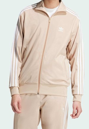 Mężczyzna w beżowym dresie Adidas z białymi paskami na rękawach i spodniach, stojący na jasnym tle.