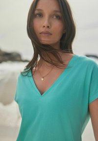T-shirt teal à col en V, manches courtes, tissu doux, coupe décontractée. Un collier doré avec un pendentif circulaire ajoute du détail.