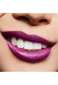 Primo piano di labbra con rossetto viola brillante e ricco. I denti sono bianchi e prominenti, con pelle liscia attorno alla bocca.