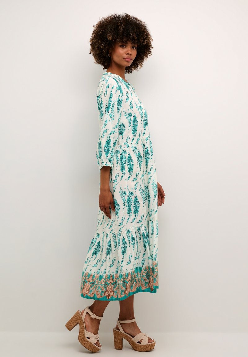 Rochie maxi cu o bază albă decorată cu modele florale în nuanțe de teal, mâneci trei sferturi și tiv volanat. Asortată cu sandale cu platformă color ciocolatiu.