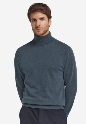 Mand med kort brunt hår iført en blå højhalset sweater og mørke bukser, stående foran en hvid baggrund.