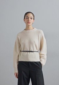 Lys beige genser med lange ermer og rund hals, brukt over svarte høyhålte bukser, med et tynt svart belte i livet.