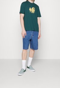 Grøn bomulds t-shirt med en gul grafik, blå denim shorts og grønternet slip-on sko iført hvide sokker.
