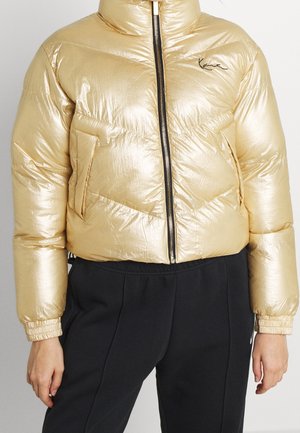 Złota kurtka puchowa o błyszczącej powierzchni, z wysokim kołnierzem, zamkiem błyskawicznym z przodu i bocznymi kieszeniami. Czerniejące akcenty na rękawach i suwaku.