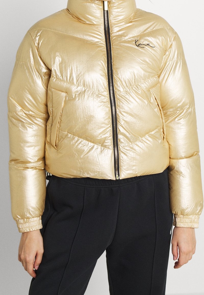 Złota kurtka puchowa o błyszczącej powierzchni, z wysokim kołnierzem, zamkiem błyskawicznym z przodu i bocznymi kieszeniami. Czerniejące akcenty na rękawach i suwaku.