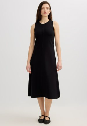 Mujer de pie con un vestido midi negro sin mangas y zapatos planos negros sobre un fondo claro y liso.