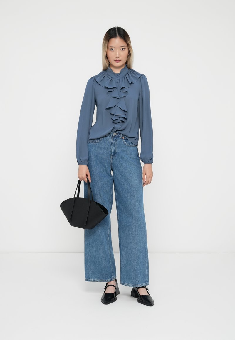 Blaue Rüschenbluse mit langen Ärmeln, kombiniert mit weiten Jeans. Das Model hält eine schwarze strukturierte Handtasche und trägt schwarze Blockabsatzschuhe.
