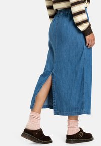 Denim maxi rok met zijsplitten, elastische tailleband, blauwe tint en gladde textuur, gedragen met donkerbruine schoenen en roze geribbelde sokken.