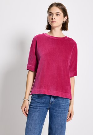 Mujer vestida con una blusa corta de manga corta de color rosa brillante y jeans azules, de pie contra un fondo claro y liso.