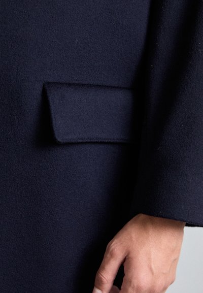 Cappotto in lana di un blu navy testurizzato, con una tasca laterale discreta e un aspetto liscio e su misura. Mano parzialmente visibile sulla manica.