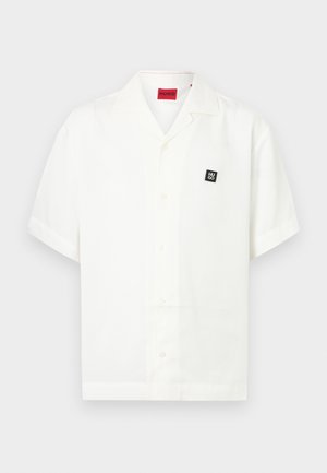 Chemise blanche à manches courtes avec col ouvert, écusson carré noir avec logo sur la poitrine et étiquette de marque rouge à l'intérieur du col.