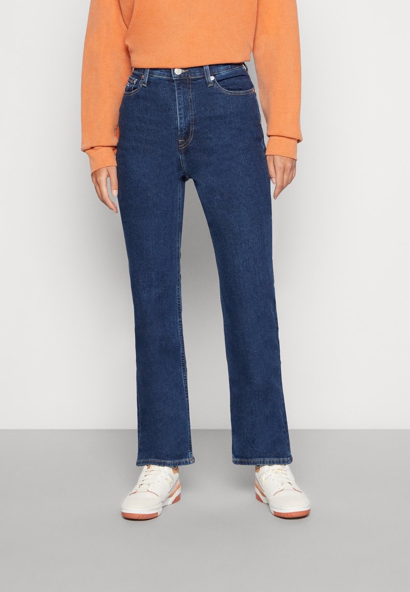 Jean en denim bleu foncé avec une coupe droite, cinq poches et une fermeture éclair. Associé à un sweat-shirt orange clair et des baskets blanches.