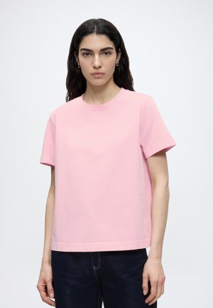 Kobieta o ciemnych włosach, ubrana w prostą, jasnoróżową koszulkę z krótkim rękawem i ciemne spodnie, stojąca na białym tle.