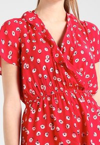 Vestido floral rojo con mangas cortas, que presenta un diseño cruzado, un cuello con volantes y patrones de flores en blanco y rosa que contrastan en una tela suave.