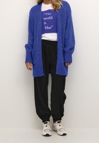 Cardigan de punto azul con frente abierto y bolsillos, usado sobre una camiseta blanca con un gráfico de texto azul, combinado con joggers negros y zapatillas.