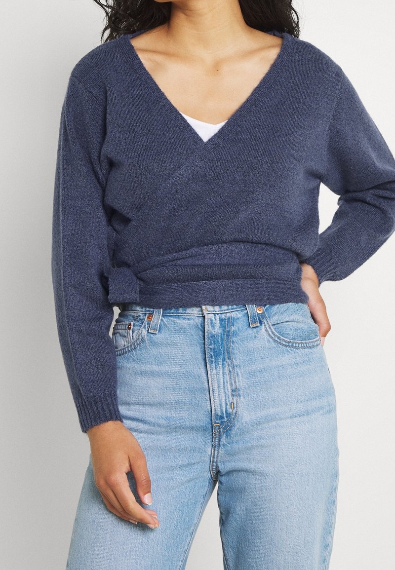 Femme portant un pull cache-cœur bleu marine sur un haut blanc avec un jean taille haute bleu clair, debout, une main sur la hanche.