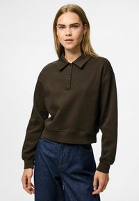 Mörkbrun tröja med polo-krage, tre svarta knappar och åtsittande ärmar. Tillsammans med mörkblå högmidjade jeans.