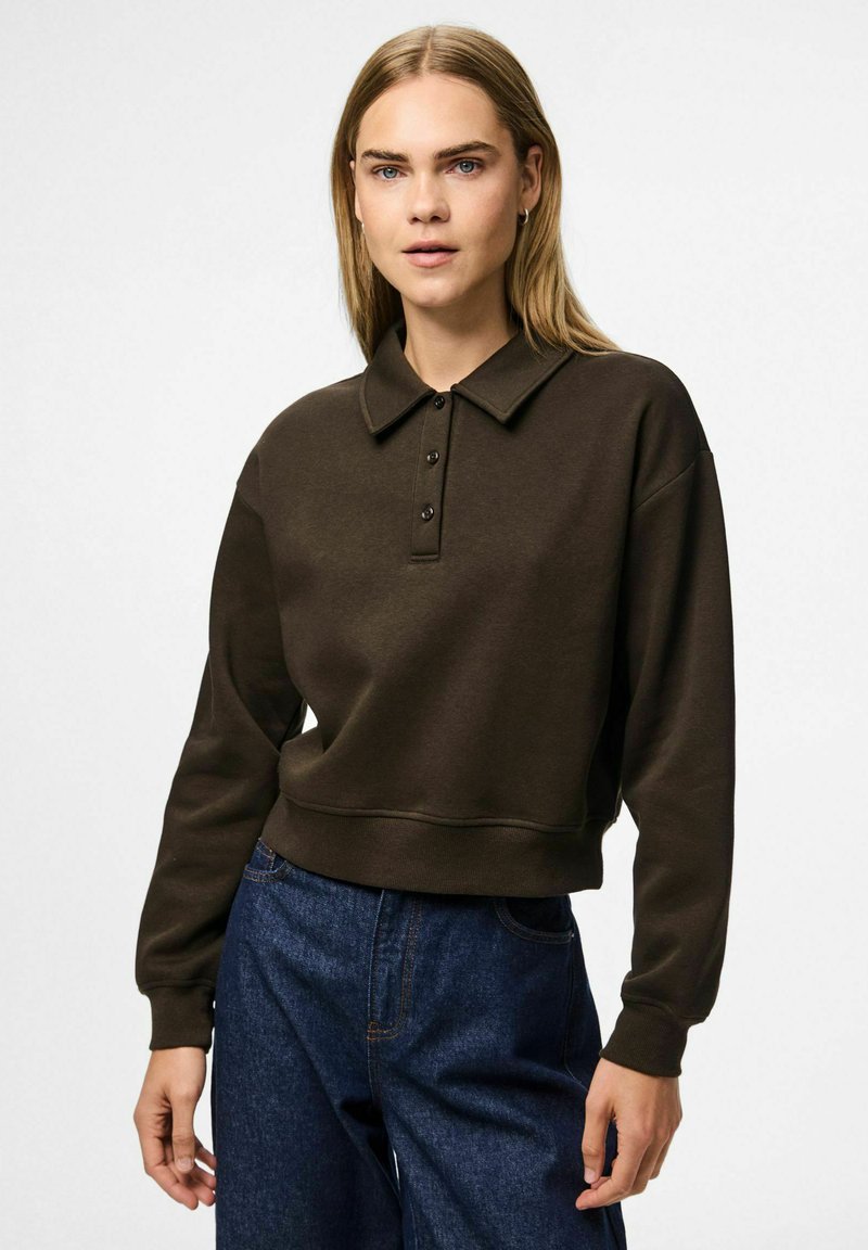 Mörkbrun tröja med polo-krage, tre svarta knappar och åtsittande ärmar. Tillsammans med mörkblå högmidjade jeans.