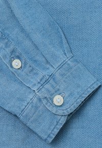 Primo piano di una camicia in denim blu, che mostra un polsino abbottonato, un tessuto testurizzato con motivo a spina di pesce e bottoni bianchi in plastica.