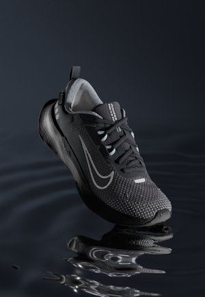 Zapatilla deportiva negra con parte superior de malla texturizada, acentos reflectantes y suela acolchada, situada sobre el agua, mostrando flexibilidad.