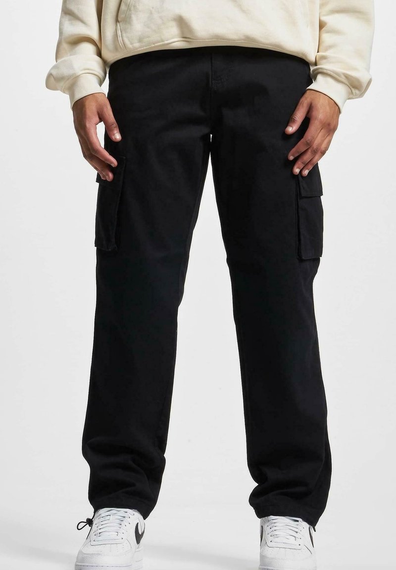 Homme portant un pantalon cargo noir avec poches latérales, des baskets blanches et un sweat-shirt de couleur claire, debout devant un fond uni.