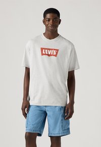 T-shirt de algodão cinza com um logo vermelho da Levi's. Modelo usa calções de ganga azuis. Design simples de gola redonda, mangas curtas, ajuste descontraído.