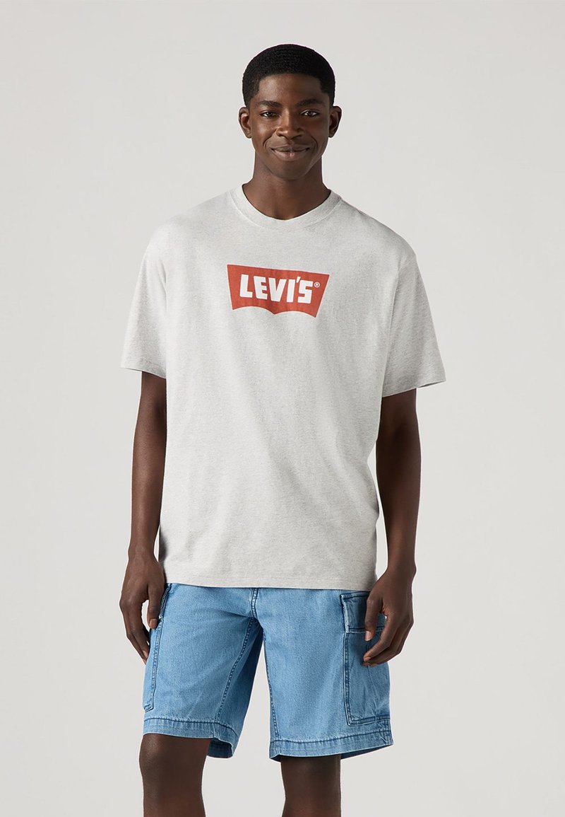 T-shirt de algodão cinza com um logo vermelho da Levi's. Modelo usa calções de ganga azuis. Design simples de gola redonda, mangas curtas, ajuste descontraído.