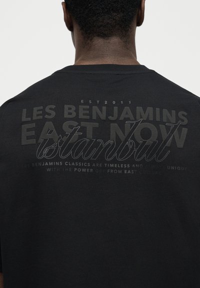 Πίσω μέρος μαύρου πουκαμίσου που φοράει ένα άτομο με κοντά μαλλιά, με μεγάλο σκούρο κείμενο που γράφει "LES BENJAMINS EAST NOW Istanbul."