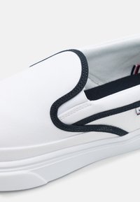 Weißer Slipper mit schwarzem Besatz und weißer Sohle, ausgestattet mit einem kleinen Stoffetikett an der Seite und elastischen Einsätzen für ein einfaches Anziehen.