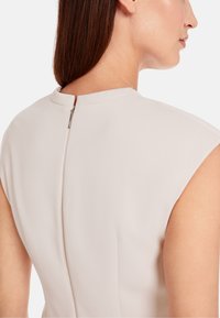 Lichtbeige jurk met een ronde halslijn, kapmouwen en een centrale ritssluiting op de rug, gemaakt van een gladde, textuurachtige stof.