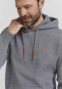 Hanorac gri în dungi cu şnururi, având accente roşii şi capete metalice. Include un logo mic pe piept. Textură de fabrică moale.