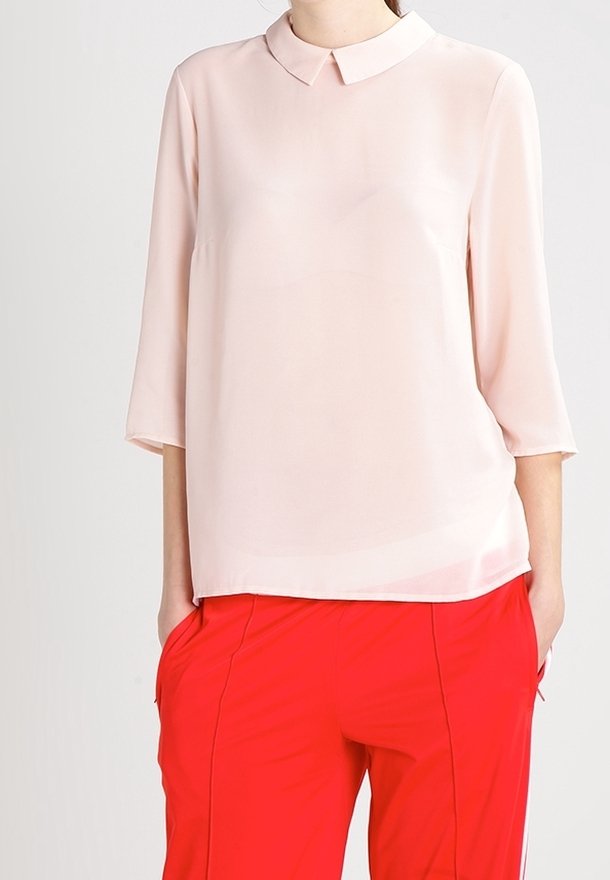 Blouse en tissu léger et transparent de couleur rose doux, dotée d'un col classique et de manches trois-quarts, associée à un pantalon rouge.