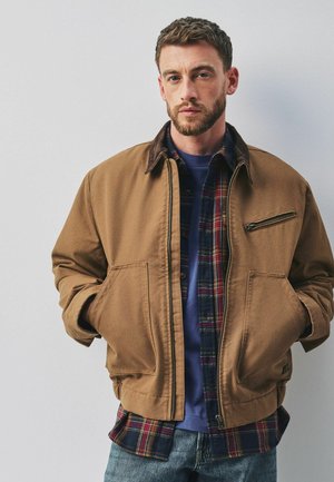 Mann mit kurzem braunem Haar und Bart, der eine beige Jacke über einem blau karierten Hemd und blauen Jeans trägt, steht vor einem einfarbigen hellen Hintergrund.