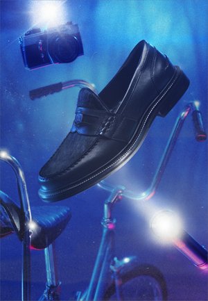 Čierna kožená loafer topánka sa vznáša nad vintage bicyklom, ktorý má riadidlá a sedlo, pričom v pozadí bliká modré svetlo fotoaparátu.