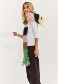 Blondynka w białej bluzce z długim rękawem, brązowych spodniach i długim, wielokolorowym, paskowanym szaliku z frędzlami na ramieniu.