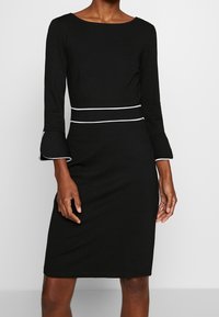 Zwarte jurk met lange mouwen en uitlopende manchetten, voorzien van een witte accentriem rond de taille en een bootnek. Glad textuur van de stof.