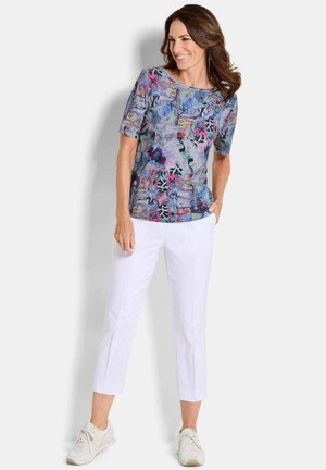 Frau lächelt, trägt ein bunt gemustertes kurzärmeliges Hemd, weiße verkürzte Hosen und weiße Sneakers, steht vor einem weißen Hintergrund.