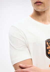 Άνδρας που φοράει λευκό κοντομάνικο πουκάμισο με πορτοκαλί και μαύρο έμβλημα λιονταριού, τα χέρια σταυρωμένα, ουδέτερο φόντο.