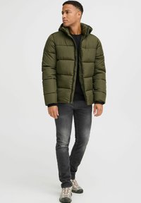 Dunjakke i mørkegrønn, quiltet design med hette, svart glidelås og sidelommer. Brukt med mørke jeans og grå joggesko.
