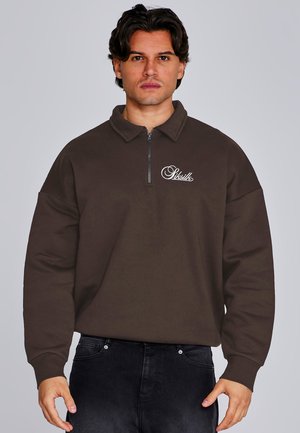 Mężczyzna w ciemnobrązowej bluzie z krótkim zamkiem i logo „Siksilk” na piersi oraz czarnych dżinsach, stojący na jednolitym szarym tle.